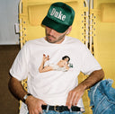 Man wearing a white t-shirt with a graphic design and a green 'Duke' cap, sitting on a yellow chair.