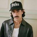 Man wearing a black cap with 'Duke' text, leather jacket, and white shirt.